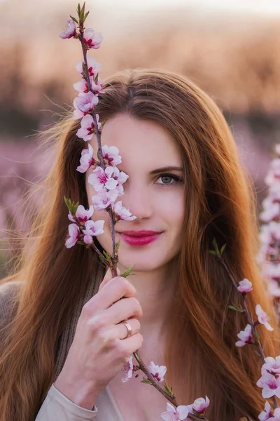 ピンクの花が咲いている枝を持つ美しい赤毛の女の子の大きな肖像画 花序は彼女の片目 赤い口紅をカバーします 女の子は カメラを見て笑顔 ソフトフォーカス — ストック写真