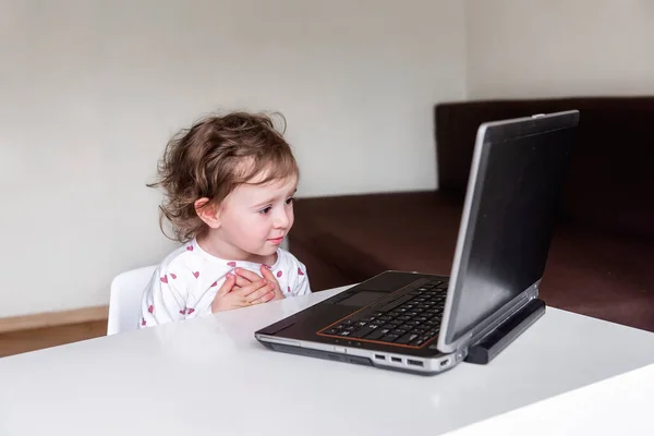 A charming little girl sits behind a laptop and watches and learns from a distance and participates in early development classes, a concept of modern early development. Selective front and back focus, artistic noises.