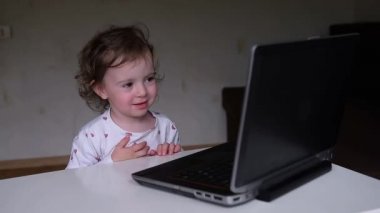Çekici küçük bir kız dizüstü bilgisayarın arkasında oturur ve uzaktan izler ve modern online anaokulu konsepti olan erken gelişim derslerine katılır. Bebek dizüstü bilgisayara bakıyor, gülüyor ve elleriyle yüzünü örtüyor.