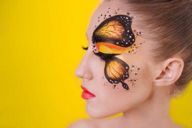 Sarı üniformalı güzel bir kız modelinin yakın plan portresi. Gözünde turuncu bir kelebek resmi, dudaklarında kırmızı ruj, topuz şeklinde sarı saçlar.. 