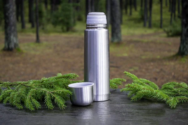 Thermos with an open lid that stands next to it. on a wooden surface with fir branches. Nature on background. The concept of outdoor recreation in the cold season.