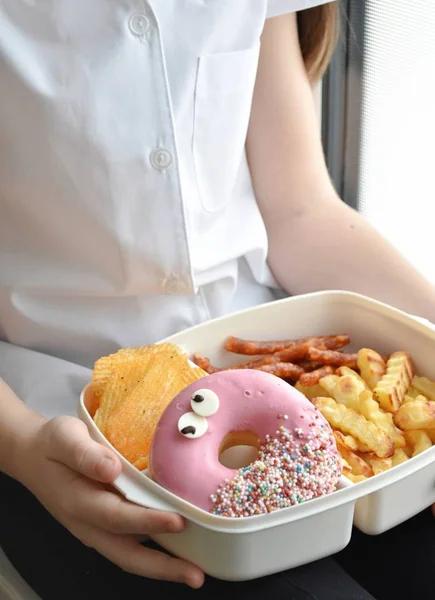 Girl holding lunchbox with unhealthy food - donut, chips, fries, sausages. Unhealthy school lunch.