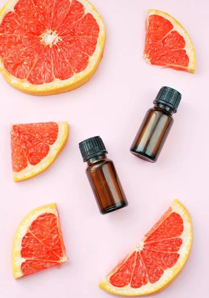 Grapefruit essential oil in dark bottles, ripe grapefruit slices on pink background flat lay.
