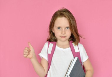 Sırt çantalı küçük tatlı bir kız. Elinde kitaplar ve pembe arka planda soyutlanmış bir parmak var. 