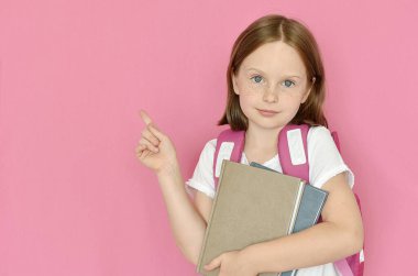 Sırt çantalı küçük tatlı bir kız. Elinde kitaplar ve pembe arka planda soyutlanmış bir parmak var. 