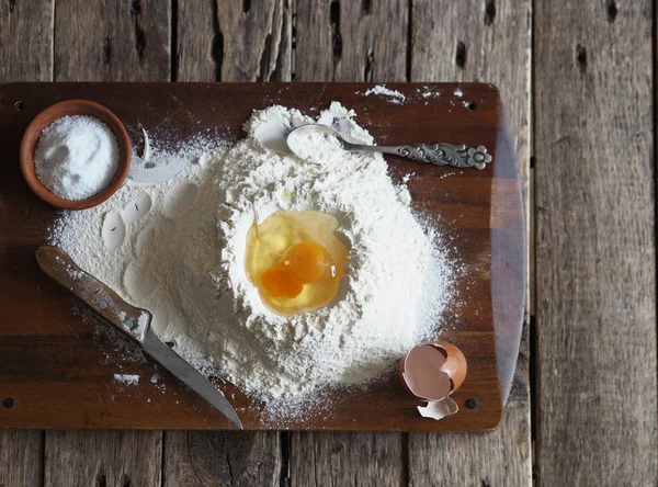 En una tabla de cortar la cocina, un huevo partido en harina con ingredientes para hacer masa. Utensilios de cocina diferentes . — Foto de Stock