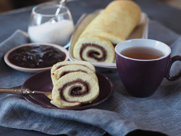 Homemade biscuit sponge roll with sweet plum jam and tea against a dark background. Natural homemade dessert. Side view.