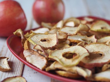 Kurutulmuş meyve ve sebzelerin faydaları. Kırmızı tabakta, beyaz ahşap bir masada kurutulmuş elma cipsi ve kırmızı taze elmalar. Organik doğal gıdalar. Yan görünüm.