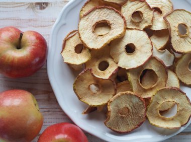 Kurutulmuş meyve ve sebzelerin faydaları. Kırmızı tabakta, beyaz ahşap bir masada kurutulmuş elma cipsi ve kırmızı taze elmalar. Organik doğal gıdalar. Üst görünüm.