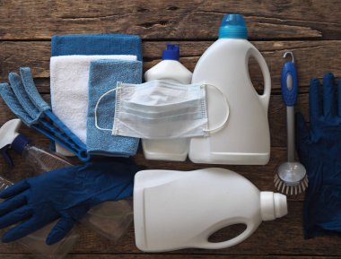 Koronavirüs. Karantina sırasında evi temizleme fikri. Doğal ahşap arka planda çevre temizleyicileri olan plastik tozluklar üzerine tıbbi koruyucu bir maske. Evin kullanımıyla evde vakit geçir..