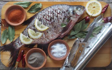 Yakın plan. Çiğ nehir balığı, fırında pişirmek için limon dilimleri, folyo rulosu ve ahşap bir tahtanın üzerinde baharat..
