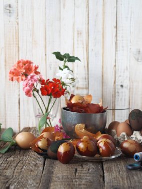 Paskalya için hazırlanıyorum. Yumurtaları yapraklarla ağ ile bağlayıp soğan kabuğunu boyarız. Pelargonium çiçekleri bir bardakta, doğal ahşap bir arka planda.