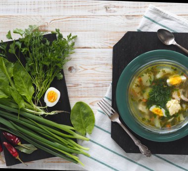 Yukarıdan manzara. Sağlıklı yiyecekler. Bir kase yeşil talaş çorbası ve beyaz ahşap bir masanın üzerindeki siyah tahtada bulunan yeşil malzemeler..