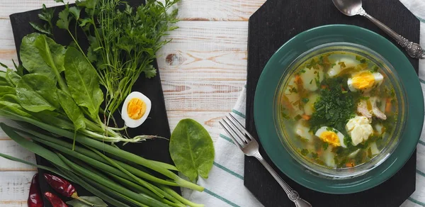 Yukarıdan manzara. Sağlıklı yiyecekler. Bir kase yeşil talaş çorbası ve beyaz ahşap bir masanın üzerindeki siyah tahtada bulunan yeşil malzemeler..