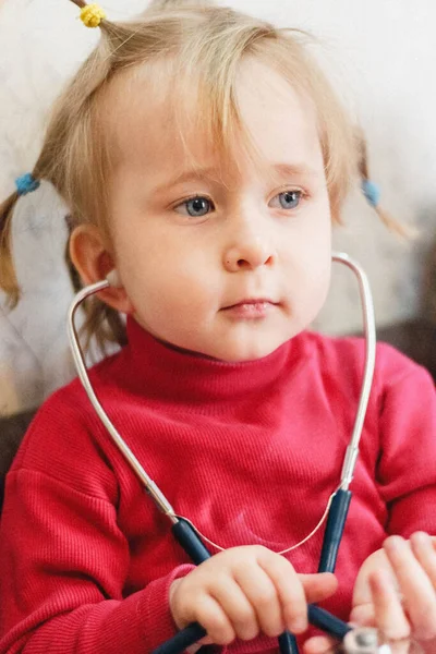 Children\'s role- playing games. Portrait of a two-year-old girl in a bright pink jacket playing doctor with a stethoscope at home.