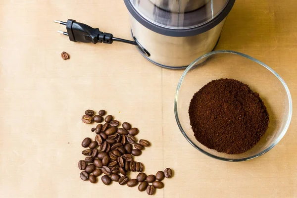 Coffee grinding at home. Electric metal coffee grinder, roasted coffee beans and ground coffee in a glass bowl on a beige table. The view from the top