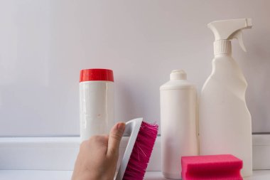 Evi temizlemek için aletler. Temizlik jeli, toz ve sprey. Beyaz plastik kaplarda temizlik ve dezenfekte etmek için. Elinde fırça ve beyaz arka planda pembe bir sünger ile..
