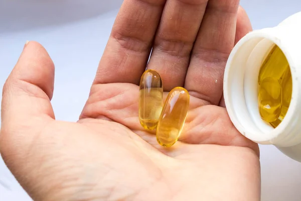 Taking food supplements and vitamins. A white can of fish oil in jelly-like transparent yellow capsules and one dose (two capsules) of capsules in a woman\'s palm close-up on a white background.