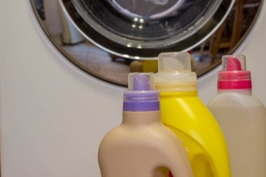 Means for washing in the washing machine. Laundry gels and rinses in bottles of different colors on the floor close-up against the background of the washing machine drum with a blurred background. clipart