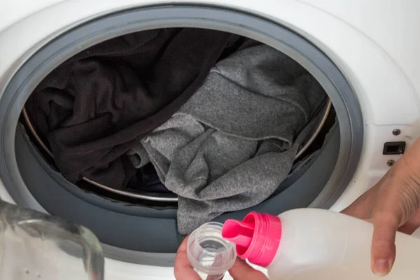 The process of preparing the washing machine for the washing cycle. A woman\'s hand pours washing gel into a measuring cap against the background of an open drum loaded with dirty clothes close-up.