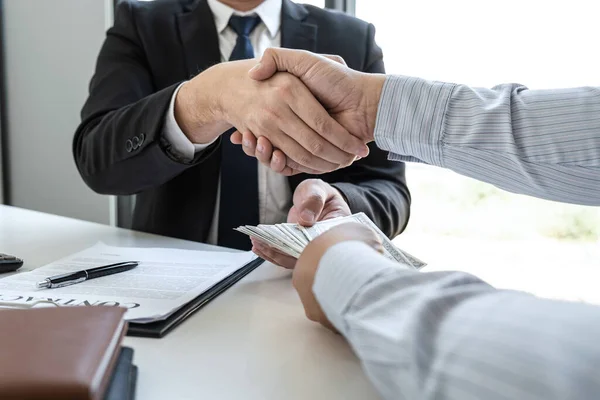 Businessman handshake with money of dollar banknotes in hands from money of their partner to give success the deal contract in a corruption scam, illegal, dishonest, bribery and corruption concept.