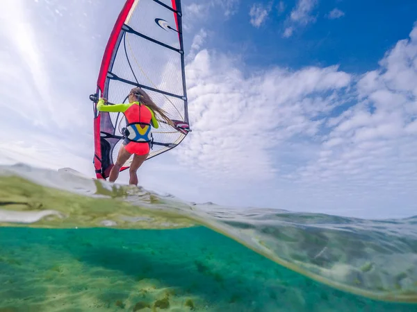 slim windsurfer girl on the board with a sail on a tropical beach, gliding on crystal clear water, view from the waterline
