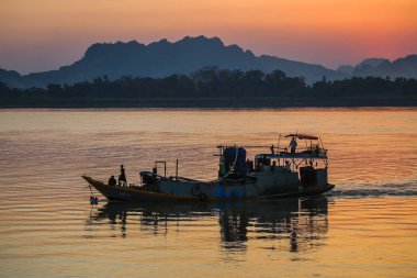 Gün batımında nehir boyunca yol alan tekne, arka plandaki dağlara karşı.