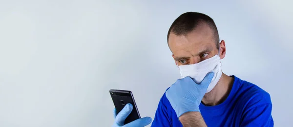 Mavi arka planda, koruyucu maskeli ve mavi elbiseli genç bir doktor. Elindeki bir cep telefonuna bakıyor. Metin için kopyalama alanı.