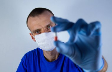 Mavi arka planda, koruyucu maskeli ve mavi elbiseli bir doktor. Elinde kırmızı bir kapsül tutuyor. Seçici odak