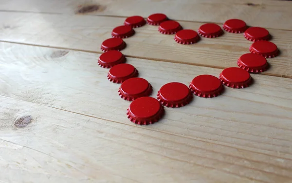 A red love heart made from beer bottle tops viewed from the side on a rustic wooden table. Beer drinkers Valentine\'s day concept. Selective focus.