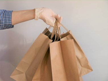 Kareli mavi gömlekli, tıbbi lateks eldivenli bir kurye Coronavirus salgını sırasında beyaz arka plandaki kahverengi kağıt poşetlerle güvenli bir şekilde satın alıyor. Kurye elinde tıbbi çantasında kahverengi kağıt poşetler tutuyor.