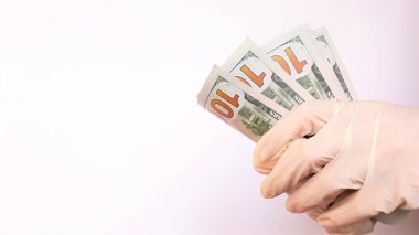 Tıbbi eldivenli eller beyaz arka planda bir tomar dolar tutuyor. Para enfeksiyonu, kirli para salgını, COVID-19, ödenmiş ilaç, tedavi ücretleri, rüşvet, yasadışı ameliyat.