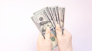 Tıbbi eldivenli eller beyaz arka planda bir tomar dolar tutuyor. Para enfeksiyonu, kirli para salgını, COVID-19, ödenmiş ilaç, tedavi ücretleri, rüşvet, yasadışı ameliyat.
