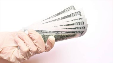 Tıbbi eldivenli eller beyaz arka planda bir tomar dolar tutuyor. Para enfeksiyonu, kirli para salgını, COVID-19, ödenmiş ilaçlar, tedavi ücretleri, rüşvetler, yasadışı ameliyatlar..