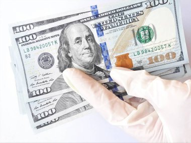 Tıbbi eldivenli eller beyaz arka planda bir tomar dolar tutuyor. Para enfeksiyonu, kirli para salgını, COVID-19, ödenmiş ilaç, tedavi ücretleri, rüşvet, yasadışı ameliyat.