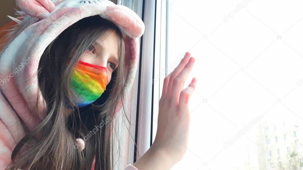 Brunette teenager girl in a medical mask painted in bright rainbow colors stands at window with her hand on glass.Concept of staying at home,staying safe.Chasetherainbow flash mob.Distance learning.