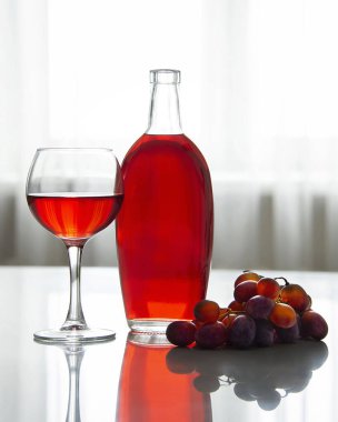 Bir şişe kırmızı şarap, bir kadeh kırmızı şarap ve pencereye dayalı beyaz bir masanın üzerinde koyu üzümler. Dikey yönelim.