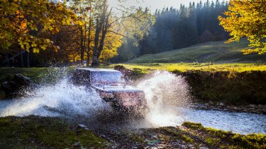 Nehir suyunun içinden geçen sıra dışı bir off-road arabası gün batımında Carpahtians Dağlarında su sıçratır ve kıvılcımlar saçar. Arka planda güzel bir güneş ışığı vardır.