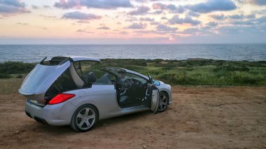 Çatı mekanizması günbatımına karşı çalışan üstü açık gri bir araba. Spor Cabriolet otomobili deniz kıyısındaki bir adanın tepesine iniyor.