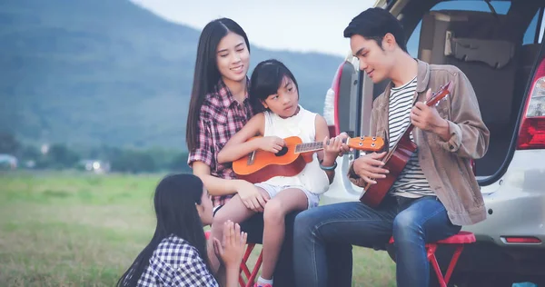 Lycklig liten flicka spela ukulele med asiatisk familj som sitter i t — Stockfoto