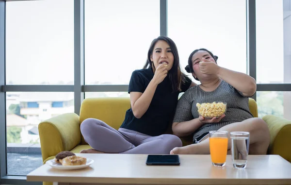 Mujer con sobrepeso y chica asiática disfrutan comiendo comida en el sofá en casa — Foto de Stock