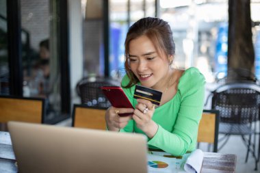 Asyalı kadınlar kafede kahve içip internetten alışveriş yapıyorlar ve akıllı telefonlardan kredi kartıyla ödeme yapıyorlar.