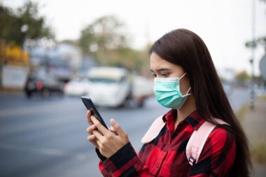 Şehirde akıllı telefon kullanan Asyalı genç kadın hava kirliliği, partiküller ve grip virüsü, grip virüsü, grip ve koronavirüs yüzünden maske takıyor.