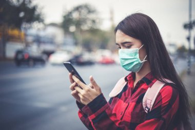 Şehirde akıllı telefon kullanan Asyalı genç kadın hava kirliliği, partiküller ve grip virüsü, grip virüsü, grip ve koronavirüs yüzünden maske takıyor.