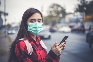 Şehirde akıllı telefon kullanan Asyalı genç kadın hava kirliliği, partiküller ve grip virüsü, grip virüsü, grip ve koronavirüs yüzünden maske takıyor.