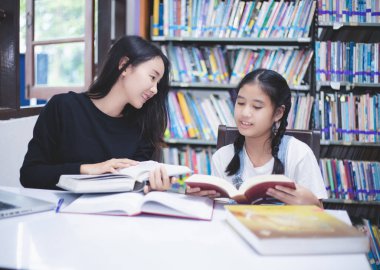 İki Asyalı kız öğrenci kitap okuyor ve kütüphanede defter kullanıyorlar..