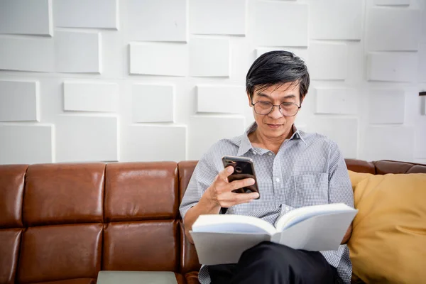 Старший Азіатський Чоловік Який Читає Книжку Користується Розумним Телефоном Дивані — стокове фото
