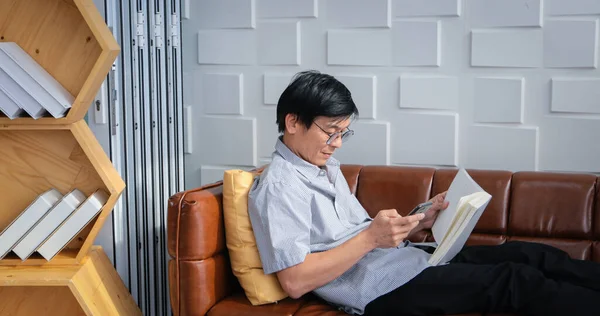 本を読んで自宅のリビングルームでソファの上でスマートフォンを使用しているシニアアジアの男性アジアの高齢者の肖像画は 雑誌を読むとリラックスして幸せです — ストック写真