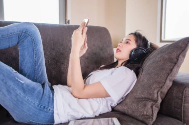 Asyalı, mutlu, neşeli, güzel bir kadın cep telefonundan müzik dinlemek için kulaklık takıyor ve akıllı telefon çalıyor ve evdeki kanepede uzanıyor., 