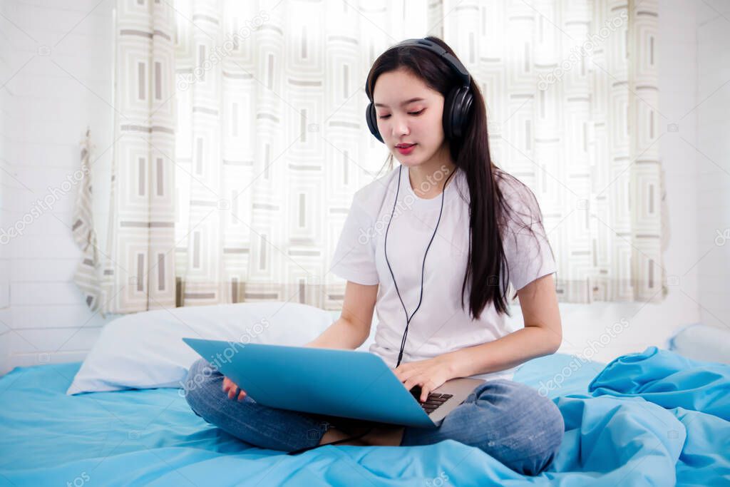 Asian happy young woman wear headphones and listening music song and watching website for shopping online from laptop  on bed at home